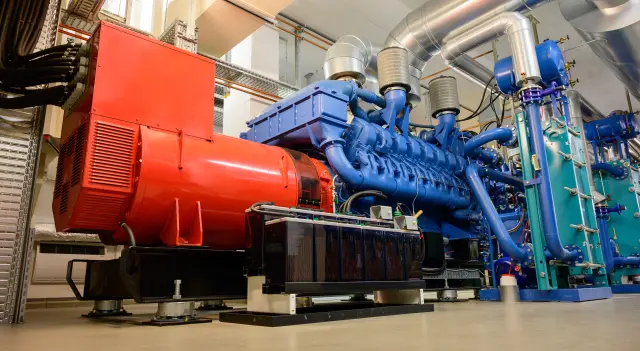 Große industrielle Generatoranlage mit blauem Motor, rotem Generatorgehäuse und angeschlossenen Lüftungs- sowie Rohrsystemen in einem Technikraum.